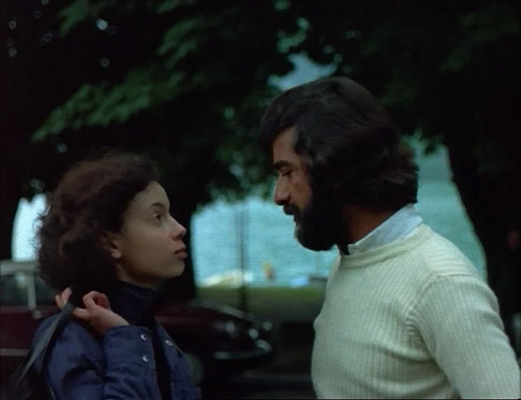 Claire’s Knee (1970) movie still: medium — A man and woman stand near a tree with a blurred body of water behind. They face each other while engaged…