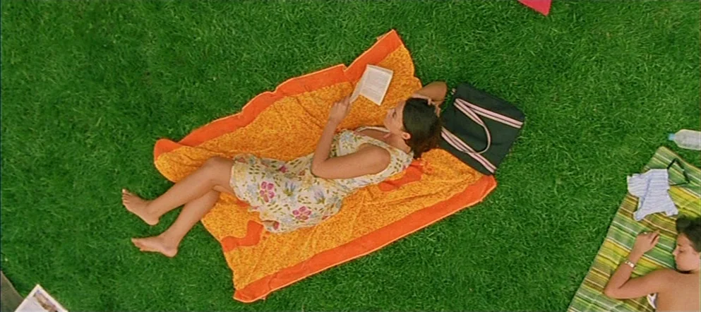 Irréversible (2002) movie still: wide — A vibrant green lawn holds two people relaxing on colorful blankets under the sun. A woman lies on an orange…