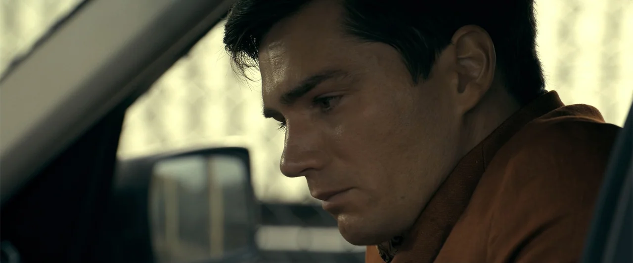 The Beekeeper (2024) movie still: closeup — The interior of a car during the day with a wire fence visible outside. A man in a brown jacket looks…