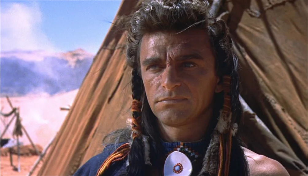 The Searchers movie still: closeup — A dry, dusty encampment with brown canvas tents set against a hazy desert background. A man with dark braided…