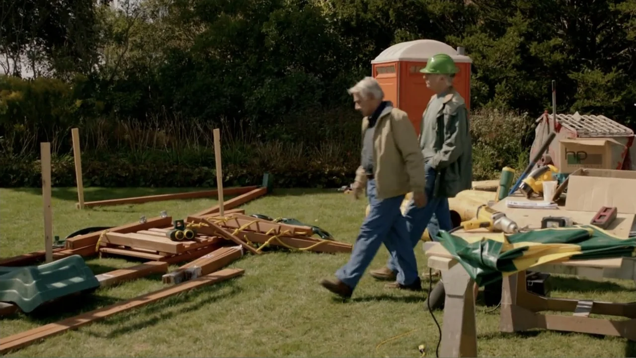 Clear History (2013) movie still: fullbody — A sunny backyard features scattered wooden beams, tools, and a portable toilet. Two men in work…