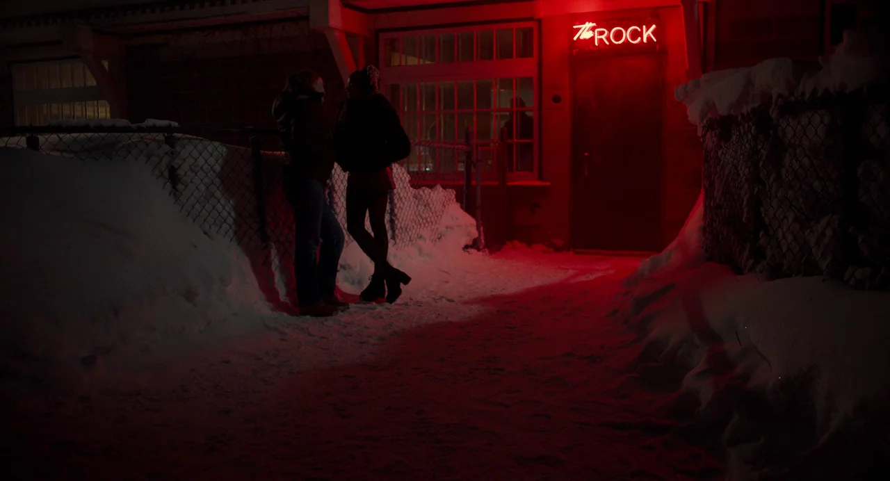 My Animal (2023) movie still: fullbody — A snow-covered path leads to a dark doorway marked by a red neon sign. Two individuals stand in the snow…