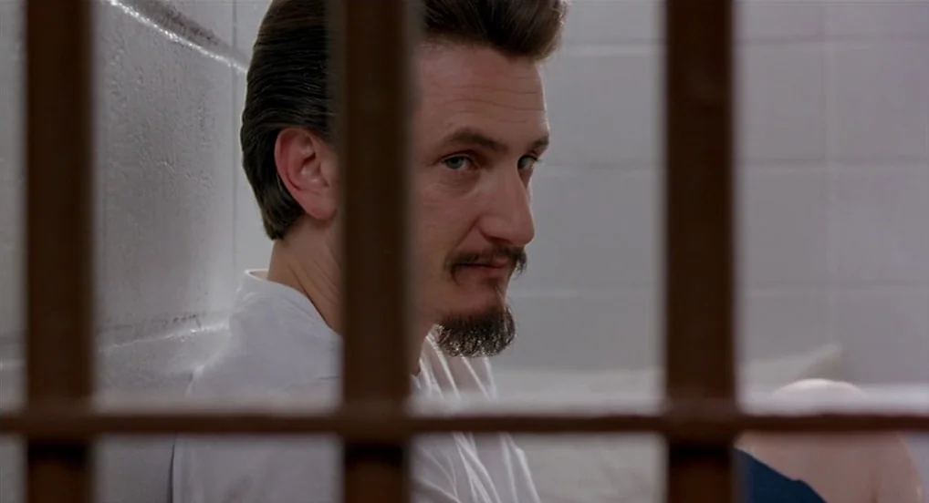 Dead Man Walking (1995) movie still: closeup — A small white-walled room enclosed by vertical metal bars in the foreground. A man with a goatee…