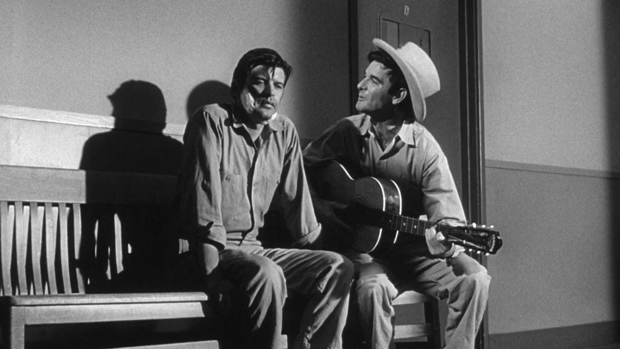 Shock Corridor (1963) movie still: medium — A man sits on a wooden bench while another man plays an acoustic guitar. The wall behind them is plain…