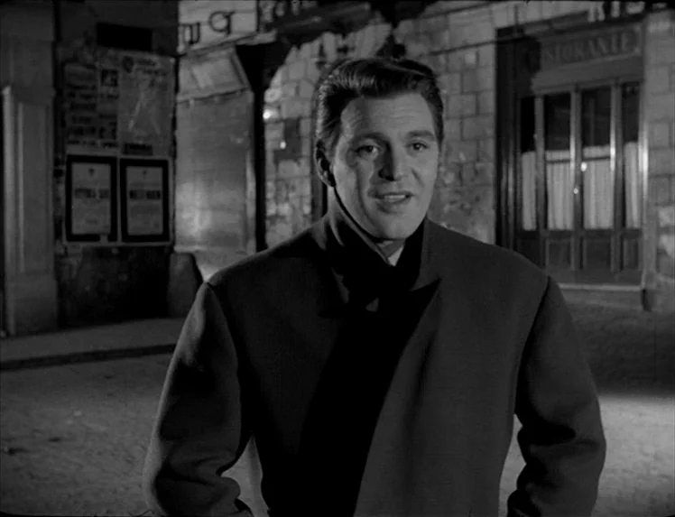 I Vitelloni (1953) movie still: medium — A night scene on a city street featuring posters on a masonry wall. The man stands facing the camera in…