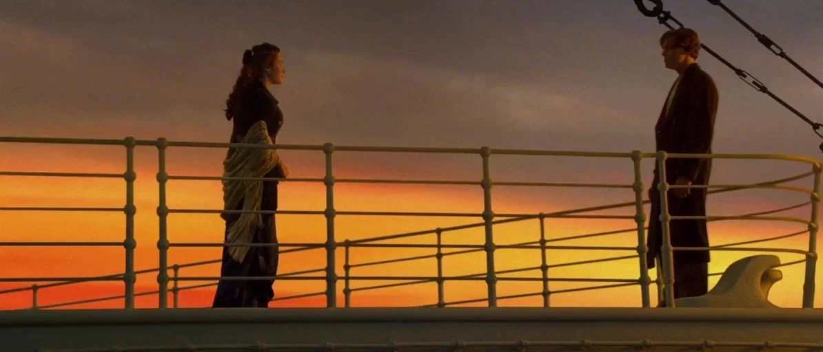 Titanic (1997) movie still: wide — A metal railing separates two individuals on the outer deck of a large ship. The sky behind them glows with…