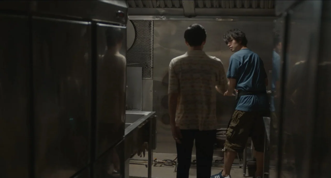 Ramen Shop (2018) movie still: medium — A narrow stainless steel food service area with metal equipment and dim overhead lighting. Two men stand by…