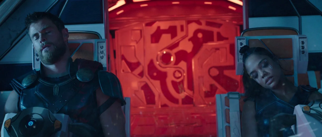 Thor: Ragnarok (2017) movie still: medium — Two individuals sit in padded ergonomic seats inside a glowing red-lit cabin. They are asleep in their…