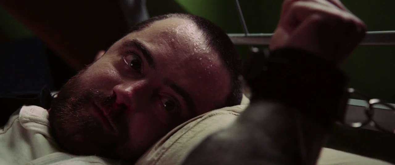 The Mind’s Eye (2015) movie still: closeup — A man lies on a hospital bed with his wrist shackled to a frame. Beads of sweat cover his forehead…