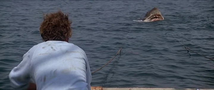 Jaws movie still: medium — A man in a light blue shirt observes the ocean surface from a boat. A large shark head emerges from the dark blue water…
