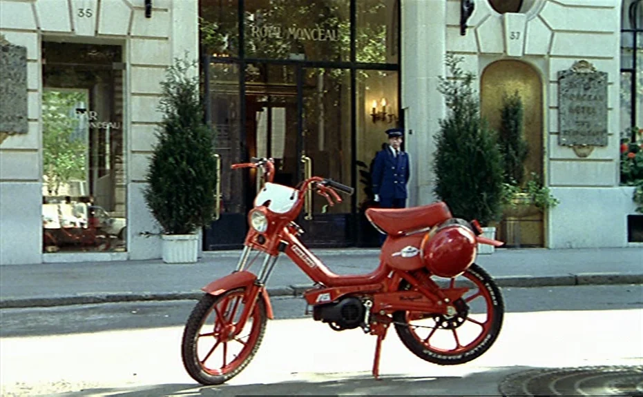 Diva (1981) movie still: medium — A bright red moped is parked on a sidewalk in front of a hotel entrance. A hotel doorman in a dark uniform stands…