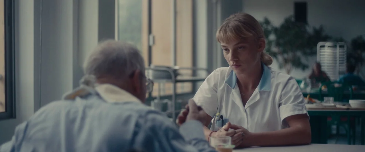 Meanwhile On Earth (2024) movie still: medium — A nursing home dining area with blurred greenery and metallic trays in the background. A young nurse…