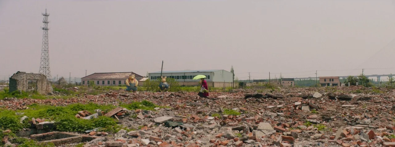 Dead Pigs (2018) movie still: wide — A vast, desolate field is covered in broken brick and concrete rubble after demolition. An elderly person walks…