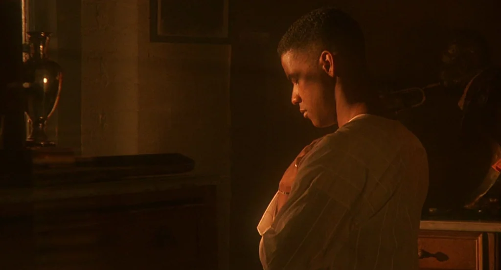 Mo’ Better Blues (1990) movie still: medium — A dimly lit room contains wooden furniture and a metallic vase on a shelf. A man stands in profile,…