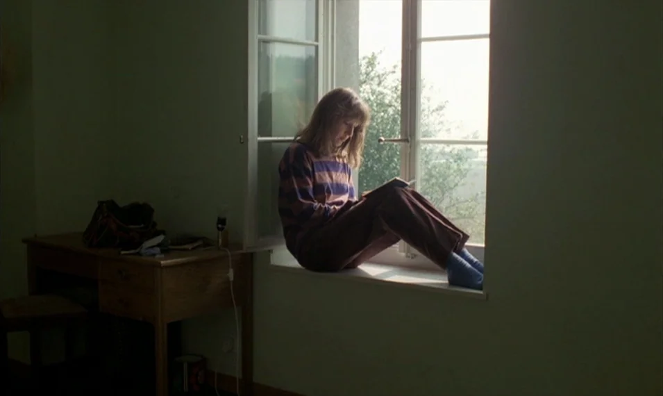 Every Man for Himself (1980) movie still: medium — A sparsely furnished room with light green walls and a wooden desk. A young woman sits on the…