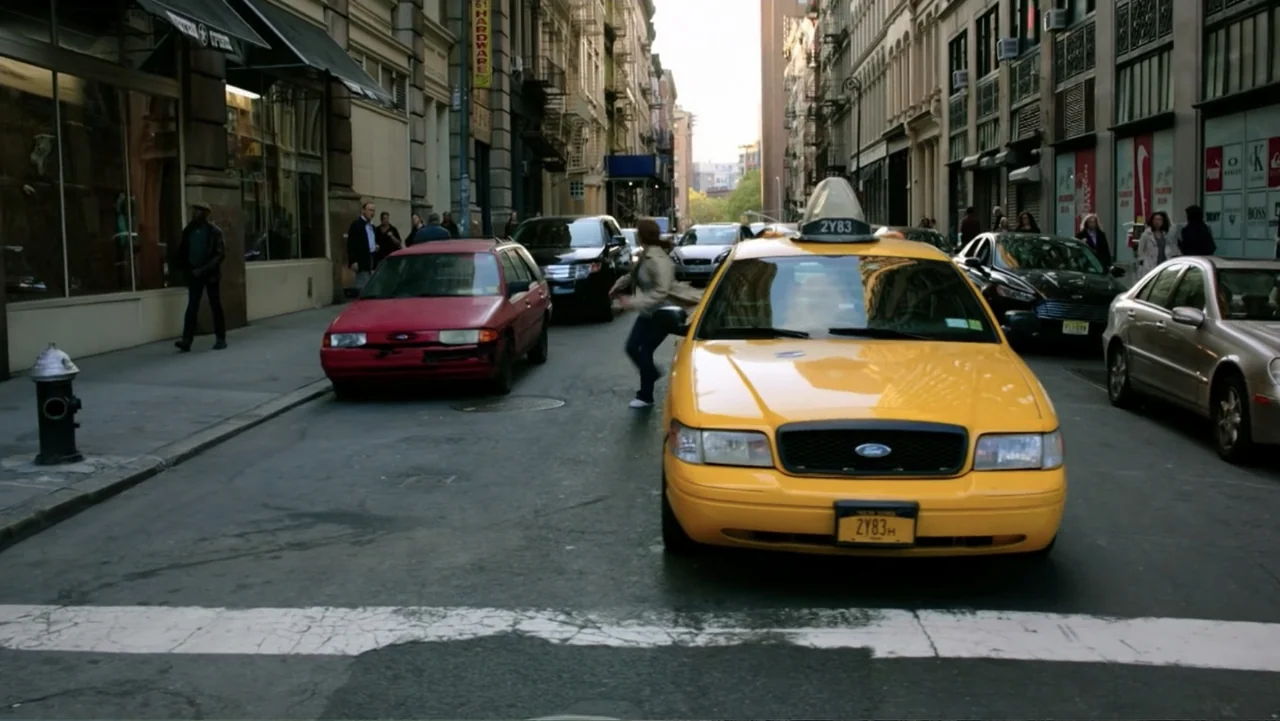 Ghostbusters (2016) movie still: wide — A narrow New York City street features multistory buildings and parked cars on both sides. A woman walks…