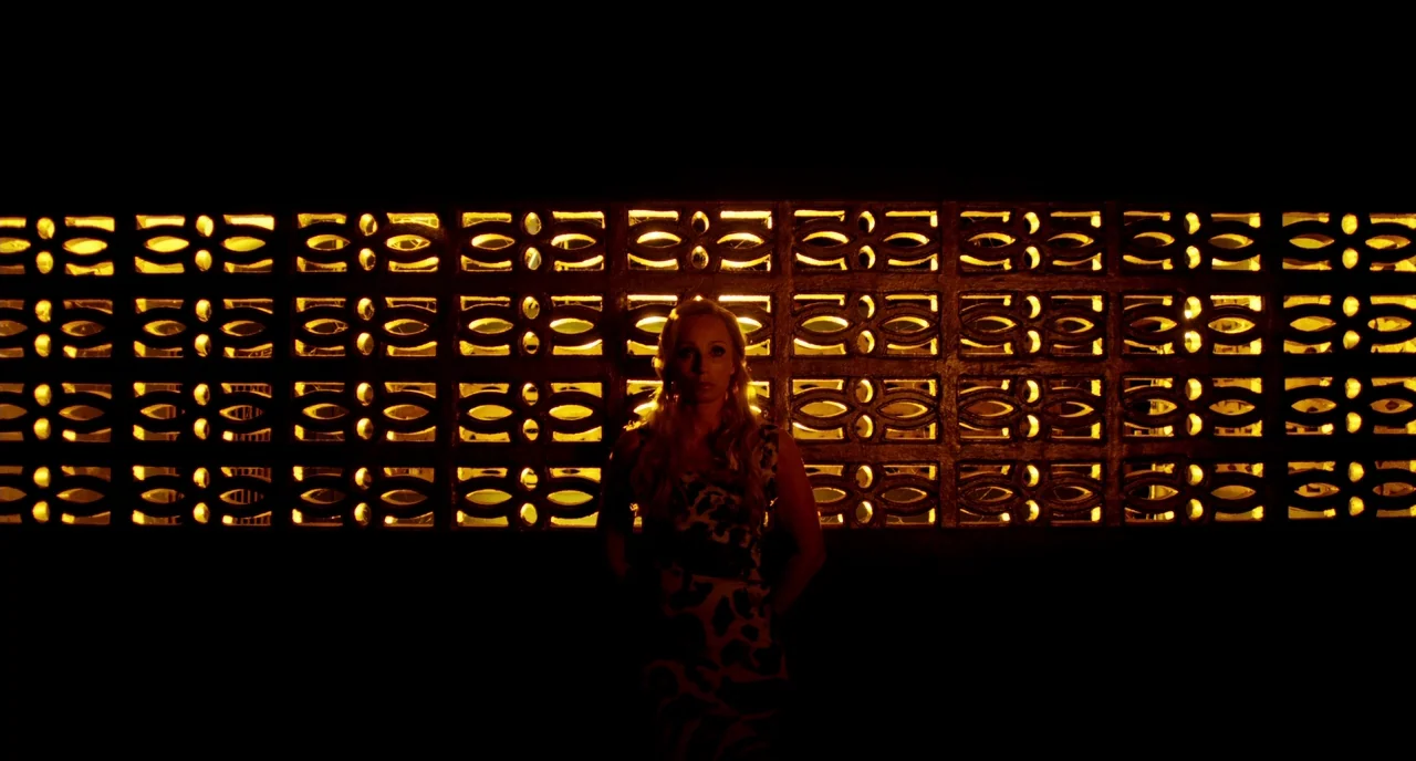 Only God Forgives (2013) movie still: medium — A woman stands centered against a backlit, decorative breeze block wall at night. The light from…