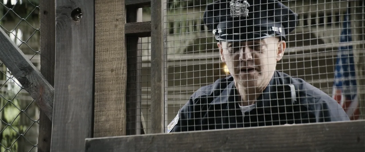 Dogman (2023) movie still: medium — A police officer stands outdoors behind a wire mesh fence and wooden frame. The man wears a dark uniform and cap…