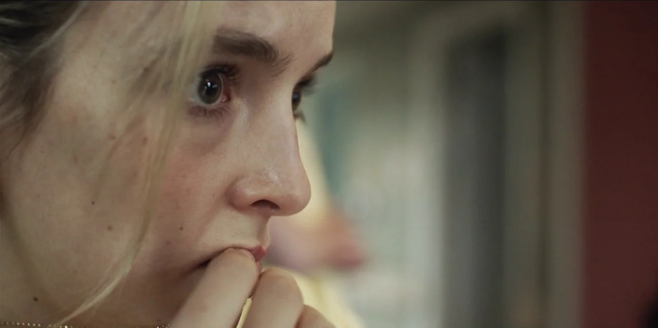Help (2021) movie still: closeup — A close-up view focuses on the face of a young woman inside a room. She looks toward the side while keeping her…