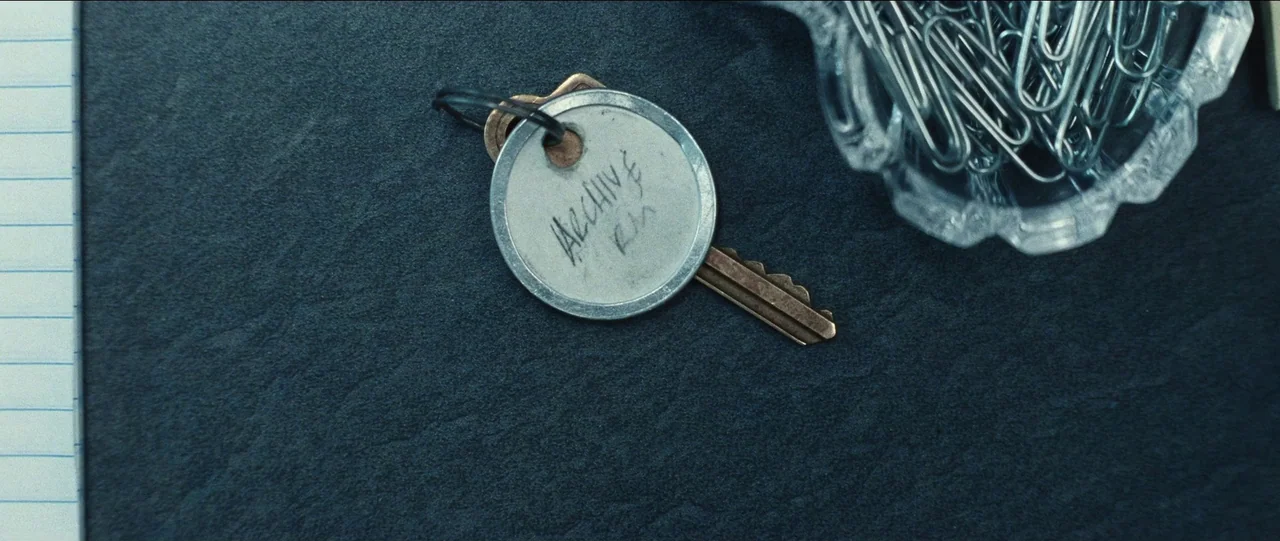 Bad Education (2019) movie still: closeup — A dark textured desktop surface holds a metal key with an attached identification tag. A small glass…