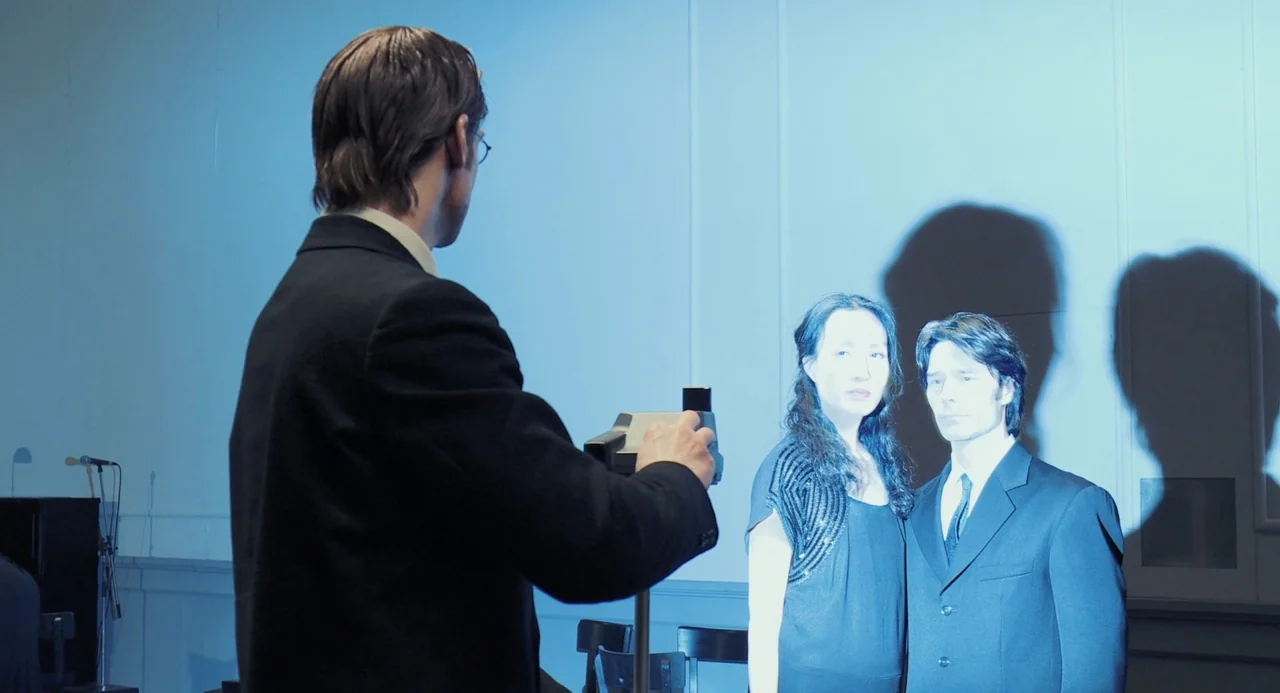 Pina (2011) movie still: medium — A man uses a digital camera to photograph a couple standing against a wall. The scene is illuminated by harsh,…