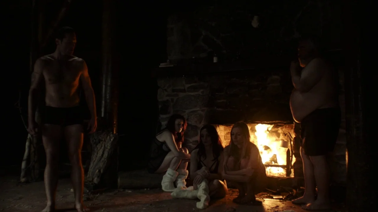 Snowbound (2017) movie still: wide — A dimly lit room features a large rustic stone fireplace emitting bright orange flames. Two men stand on…
