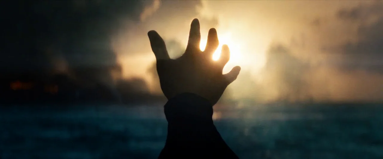 Man Of Steel (2013) movie still: closeup — A human hand reaches into the air against a bright sunset over the ocean. Smoke or mist rises from the…
