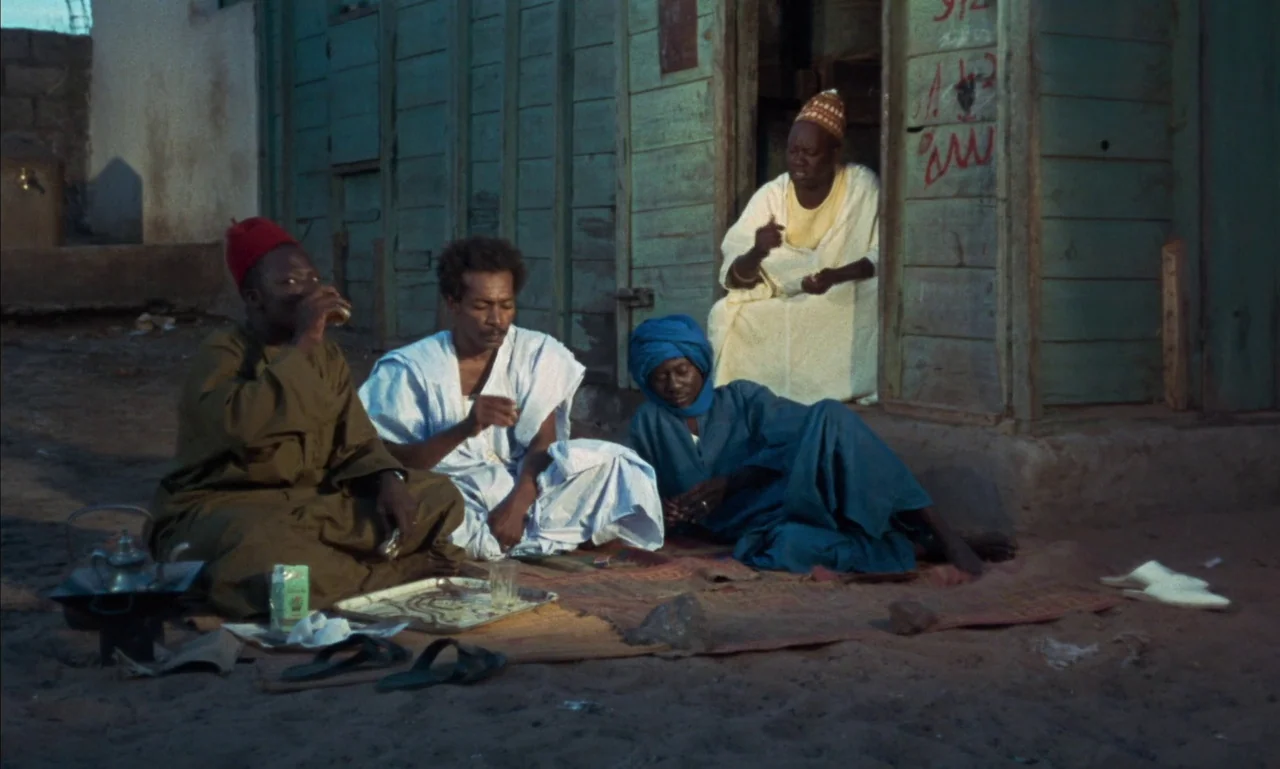 Mandabi (1968) movie still: wide — A group of men sit on a woven mat in front of a turquoise wooden shack. They drink tea together in the late…