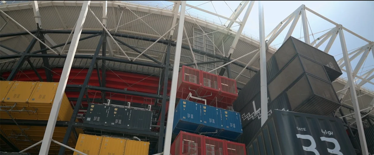 I Choose to Believe (2023) movie still: wide — A massive stadium exterior features stacked shipping containers integrated into a complex steel…