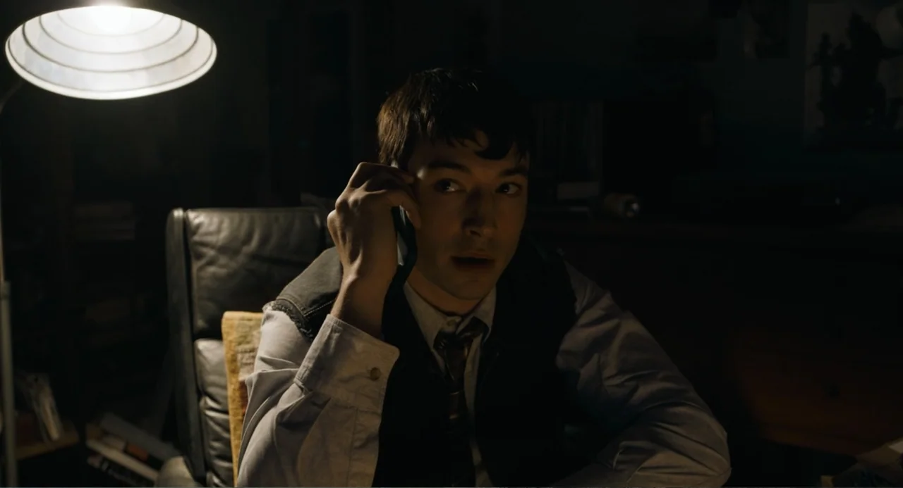 The Flash (2023) movie still: medium — A dimly lit room contains a leather chair and a desk lamp. A young man sits in the chair while speaking on a…