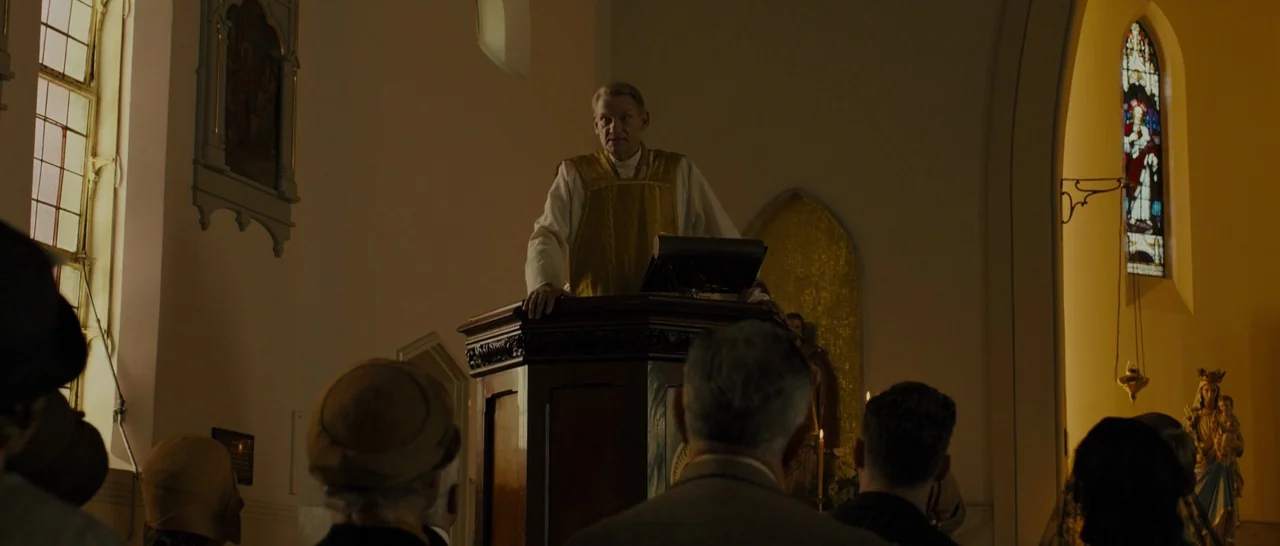 Unbroken (2014) movie still: medium — An interior of a church with rows of congregants facing a wooden pulpit. The elderly priest stands behind the…