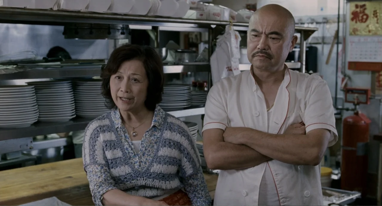 Management (2009) movie still: medium — A professional food preparation area with stainless steel shelving and stacks of plates. A man and woman…