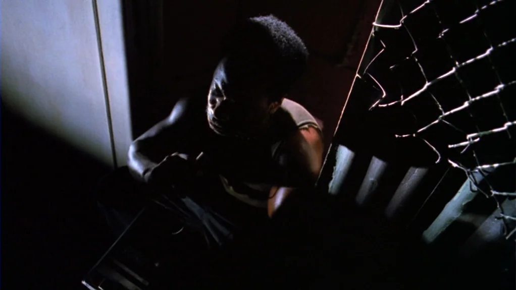 Patty Hearst (1988) movie still: medium — A dark, cramped interior stairwell featuring a metal chain-link fence along one side. A young man sits…