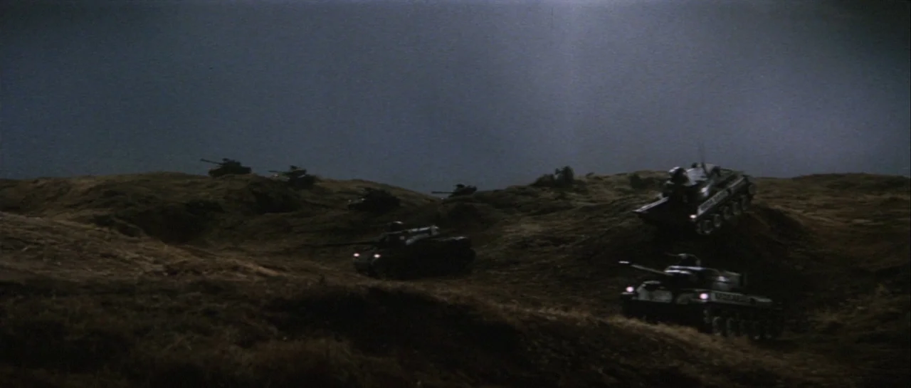 Godzilla Vs Gigan (1972) movie still: wide — A dark, hilly landscape is set against a faint, overcast night sky. Several military tanks move across…