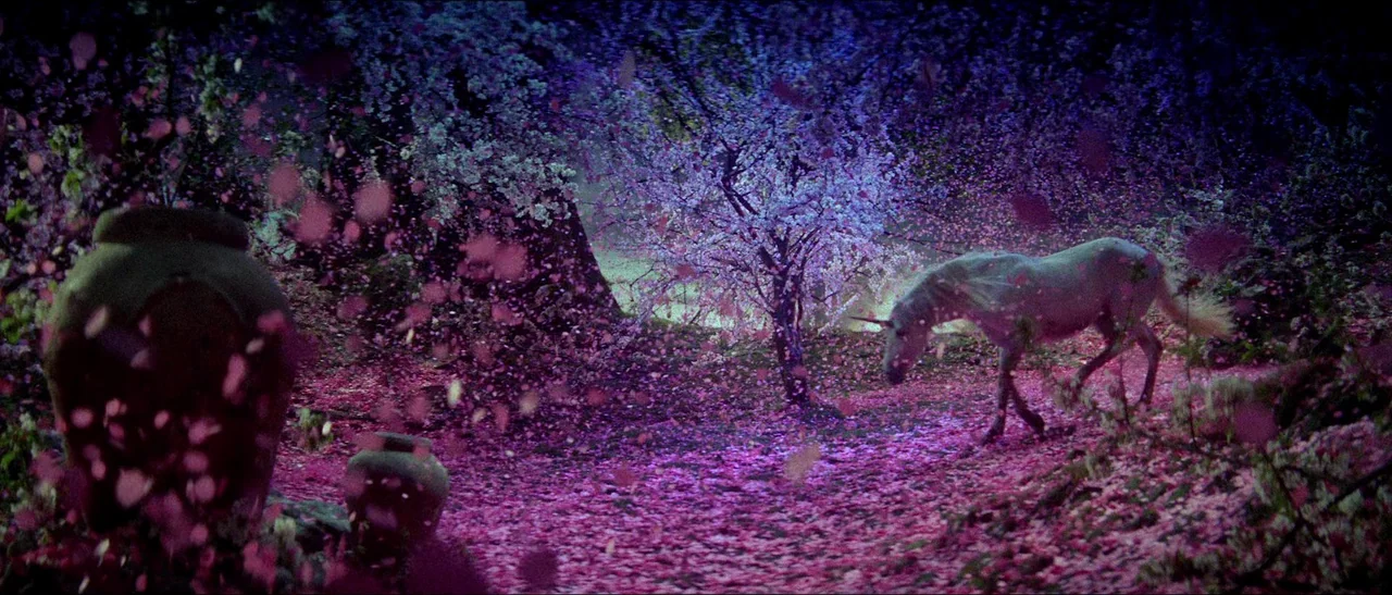 Legend (1985) movie still: wide — A dark grove is covered in thick layers of bright pink fallen flower petals. A pale white unicorn walks slowly…