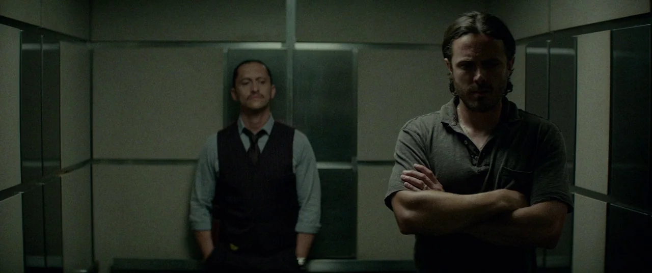 Triple 9 (2016) movie still: medium — A small elevator with paneled walls features two men standing in silence. One man in a polo shirt occupies the…