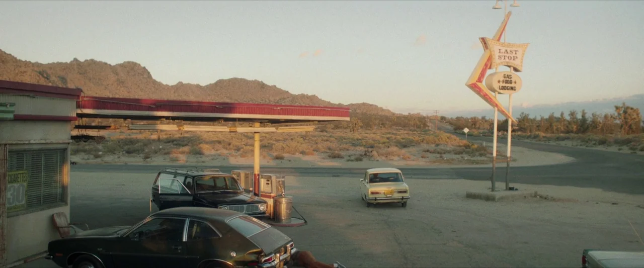 The Last Stop in Yuma County (2023) movie still: wide — A rural 1970s gas station stands in a flat desert landscape with distant mountains. Two…