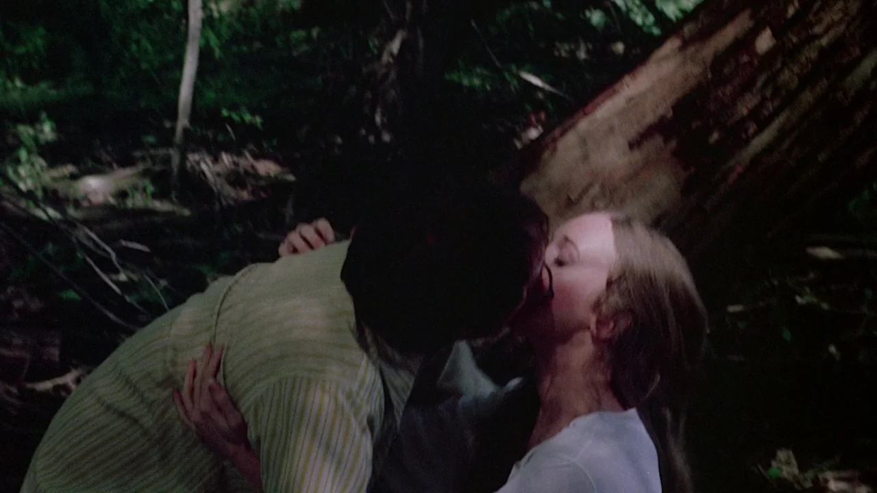 I Spit On Your Grave (1978) movie still: medium — A dense forest environment with a fallen tree trunk visible in the background. Two young people…