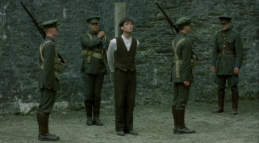 The Wind That Shakes the Barley (2006) movie still: fullbody — A man stands blindfolded and tied to a wooden stake against a stone wall. Four…