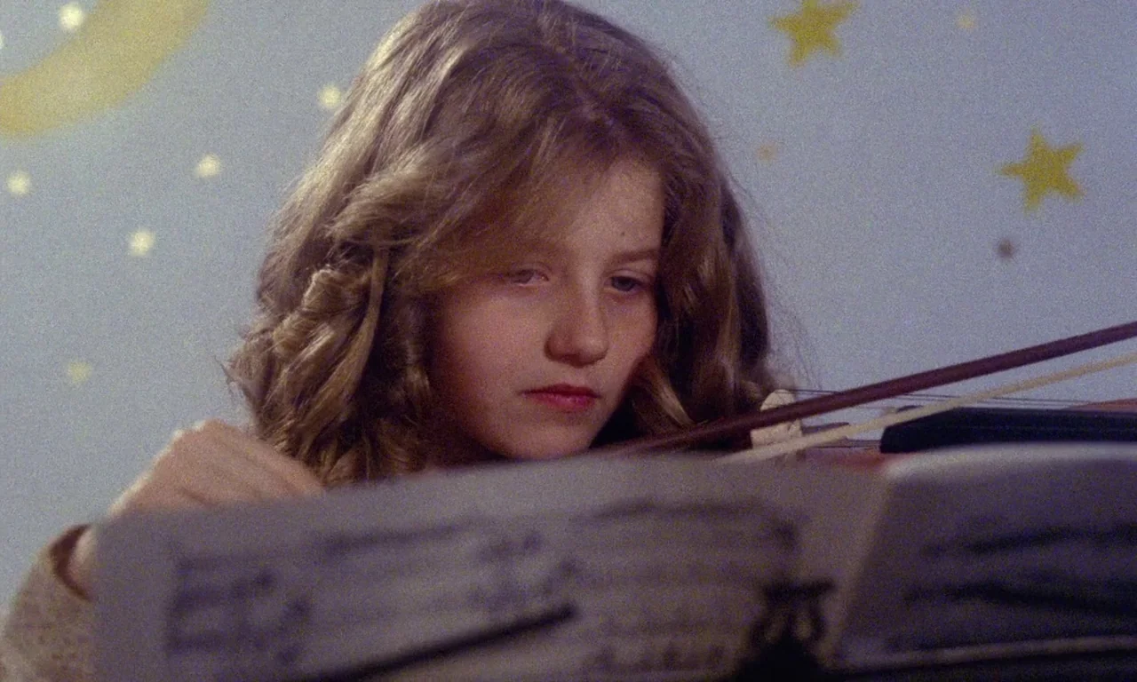 Paganini Horror (1989) movie still: closeup — A girl practices the violin in front of a wall decorated with star stickers. She stares intently at…