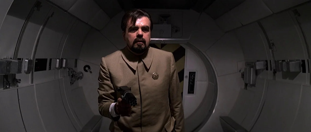 Moonraker (1979) movie still: medium — A dark, curved spaceship corridor with modular panels and a circular hatch. A man in a beige uniform stands…
