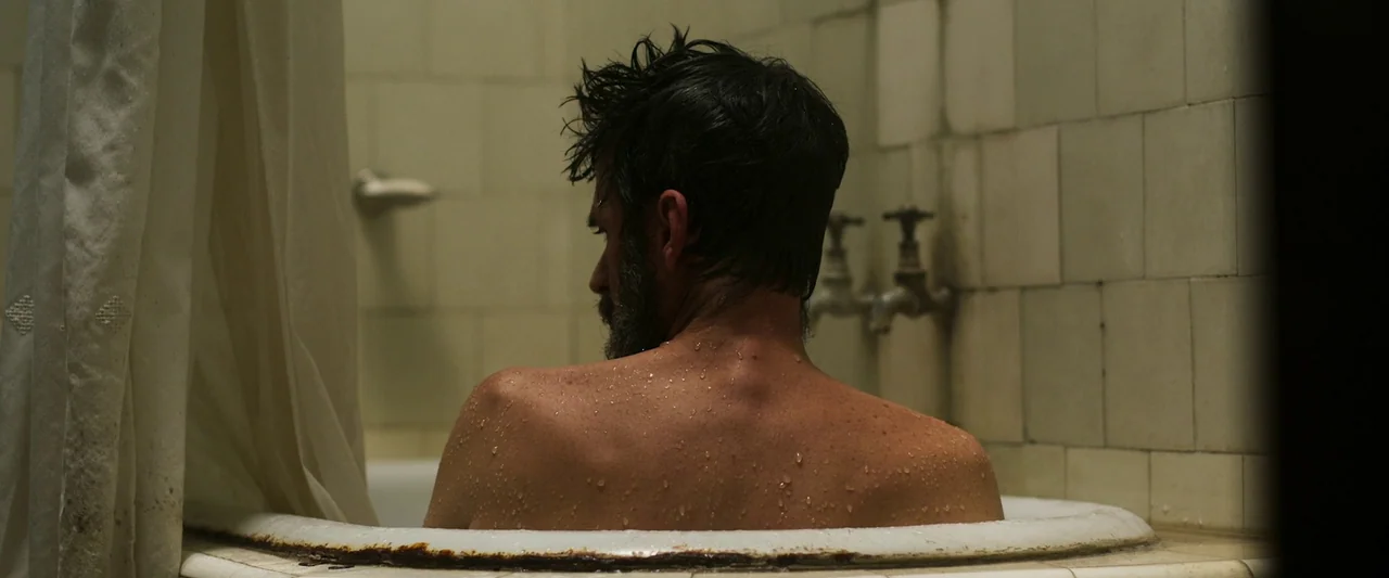 When Evil Lurks (2023) movie still: medium — A worn, aged bathtub features beige tile walls and rusted metal hardware. A man with wet hair and a…