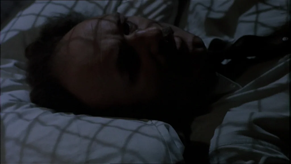 The Conversation (1974) movie still: closeup — A man lies on a bed with white linens in a darkened room. Grid patterns from a window grate are cast…
