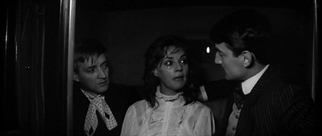 Jules and Jim (1962) movie still: medium — A dark, cramped train compartment with tight seating and wood-paneled walls. Two men flank a woman while…