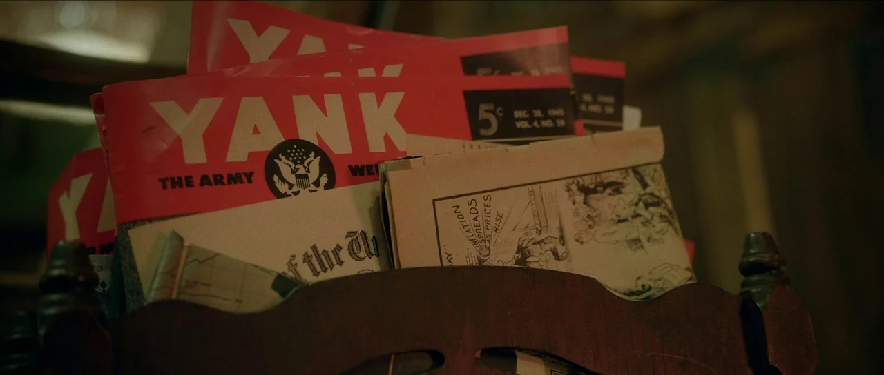 Brooklyn 45 (2023) movie still: closeup — A dark wooden rack contains several copies of Yank magazine and other papers. The magazines feature bold…
