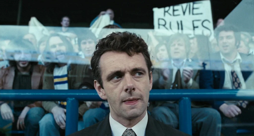 The Damned United (2009) movie still: medium — A man sits in the foreground behind a glass barrier at a sports stadium. Out-of-focus spectators in…