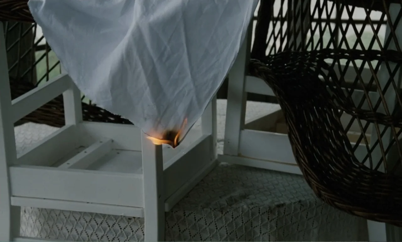 The Sacrifice (2015) movie still: medium — A white cloth draped over a white chair and wicker furniture is burning. Small orange flames consume the…