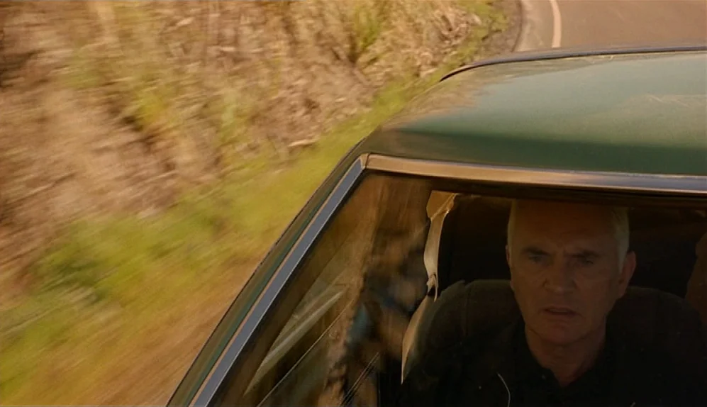 The Limey (1999) movie still: medium — An older man sits behind the wheel of a moving green car. The blurred landscape passes by rapidly outside the…
