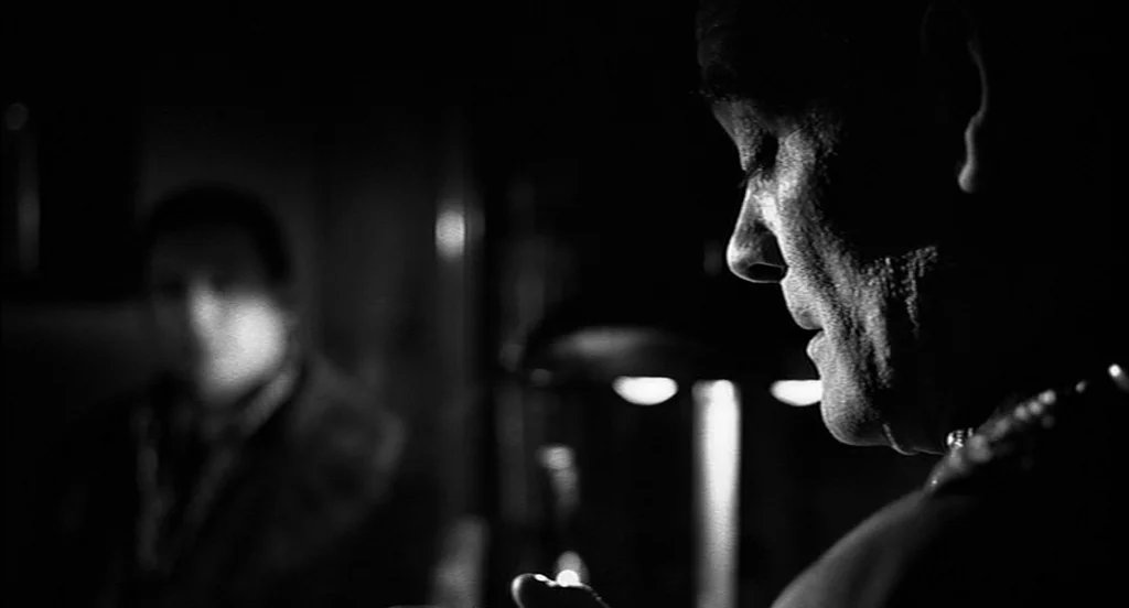 Schindler's List (1993) movie still: closeup — A dark, dimly lit room contains two figures in conversation. The man in the foreground is viewed in…