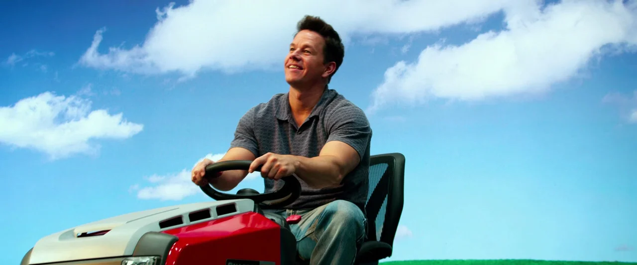 Pain & Gain (2013) movie still: medium — A man drives a red riding lawn mower against a clear, expansive blue sky. He holds the steering wheel and…
